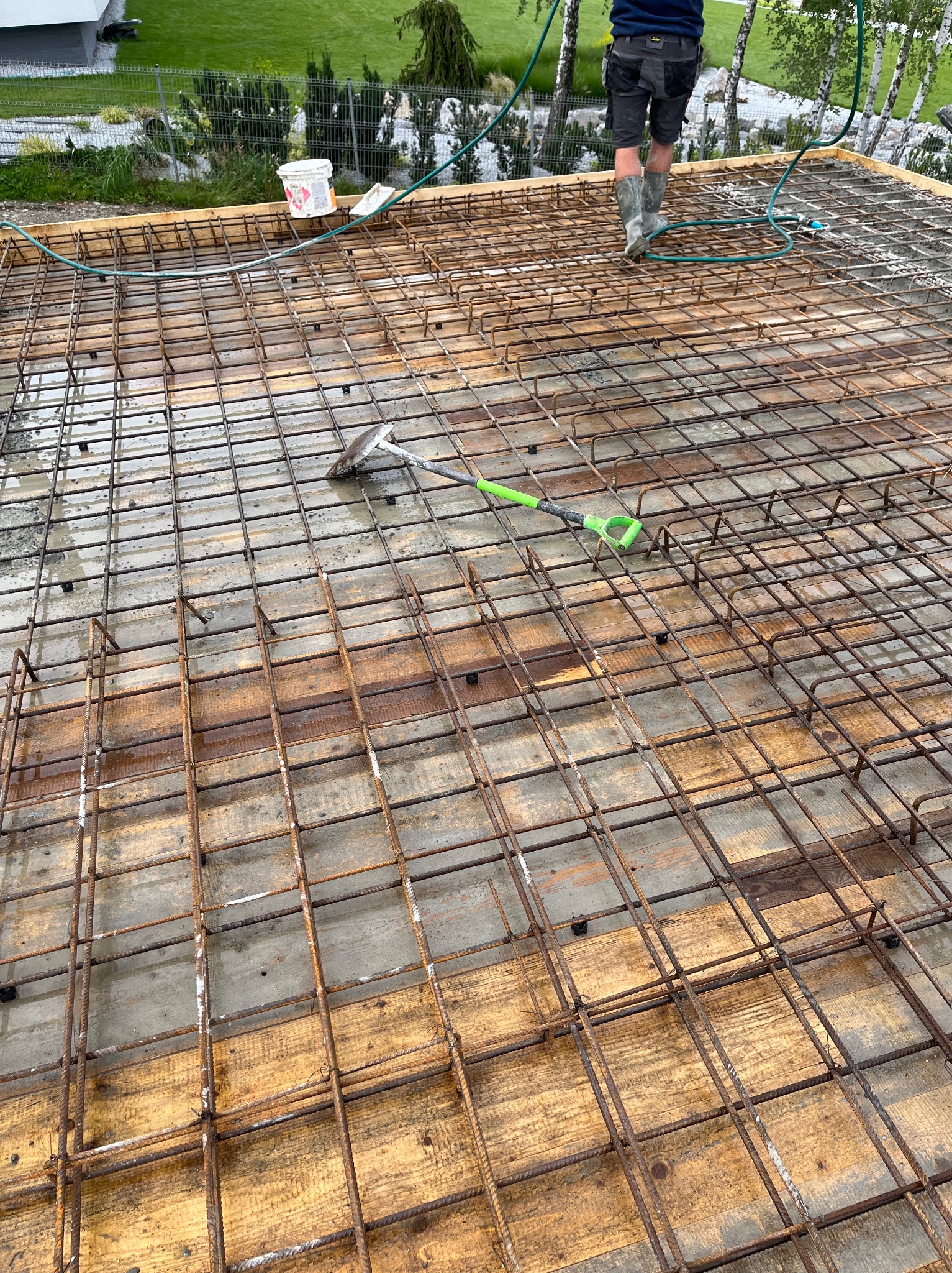 Zbrojenie płyty fundamentowej ułożone na deskowaniu z widoczną łopatą z zielonym uchwytem, pracownik w gumowych butach i wężu ogrodowym w tle.