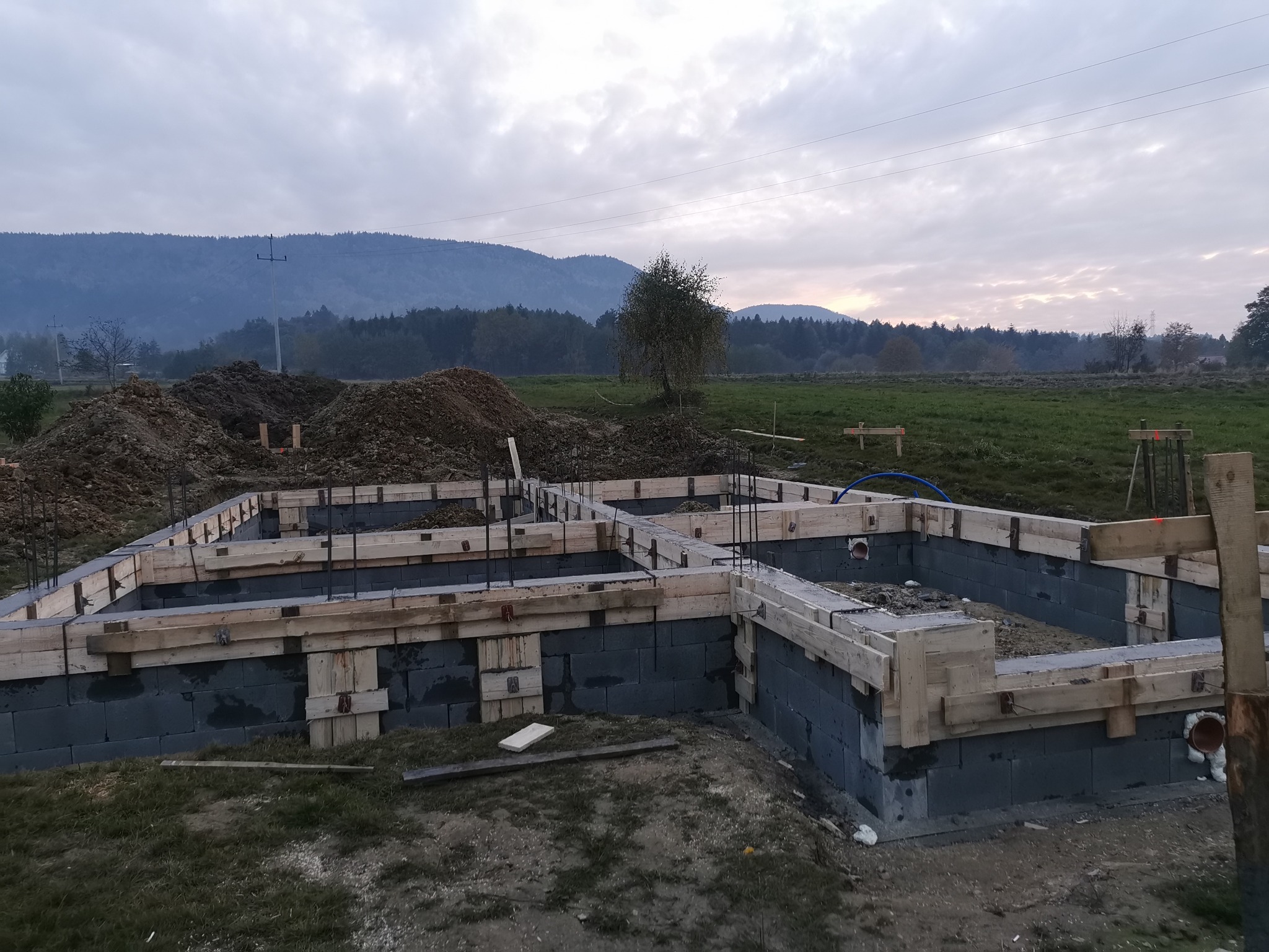 Wznoszenie fundamentów domu z bloczków betonowych, widoczne szalunki drewniane, zbrojenie pionowe, w tle hałda ziemi i zalesione wzgórza pod pochmurnym niebem.