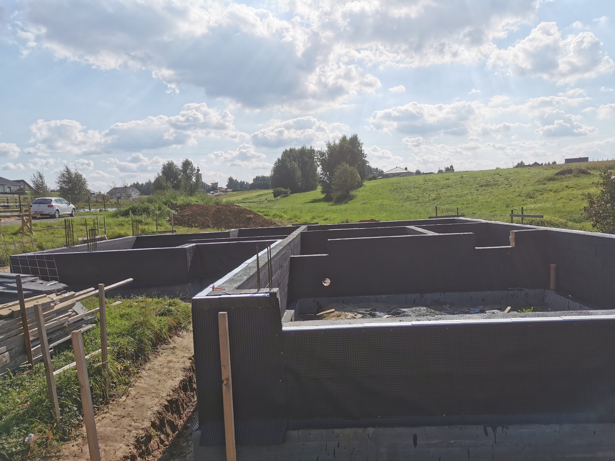 Zabezpieczone fundamenty domu jednorodzinnego z widoczną izolacją pionową, na tle zieleni i błękitnego nieba z chmurami.
