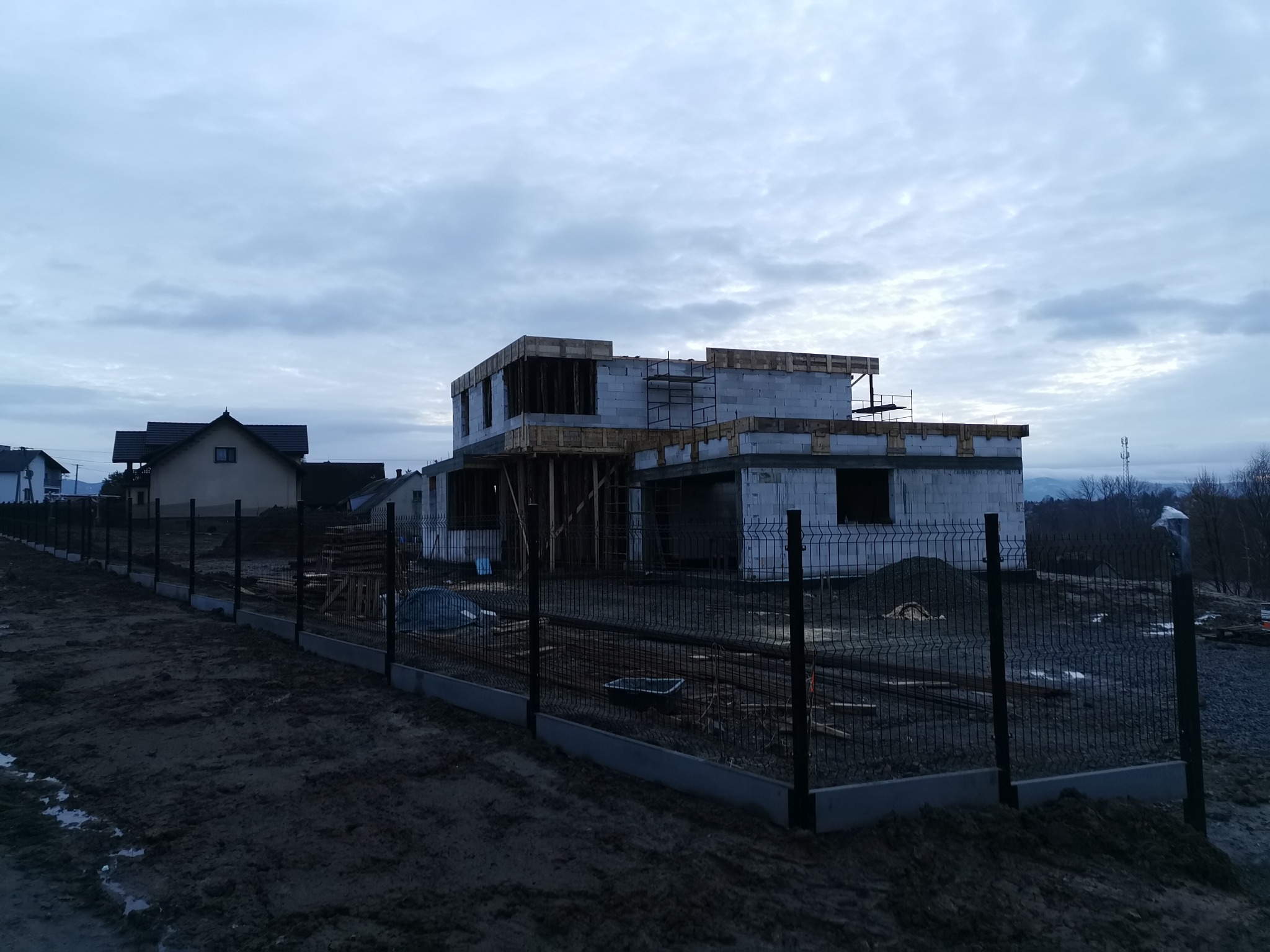 Dwukondygnacyjny dom murowany w trakcie budowy, widoczny szkielet konstrukcji z bloczków, drewniane szalunki na stropie, ogrodzenie panelowe z podmurówką, pochmurne niebo.