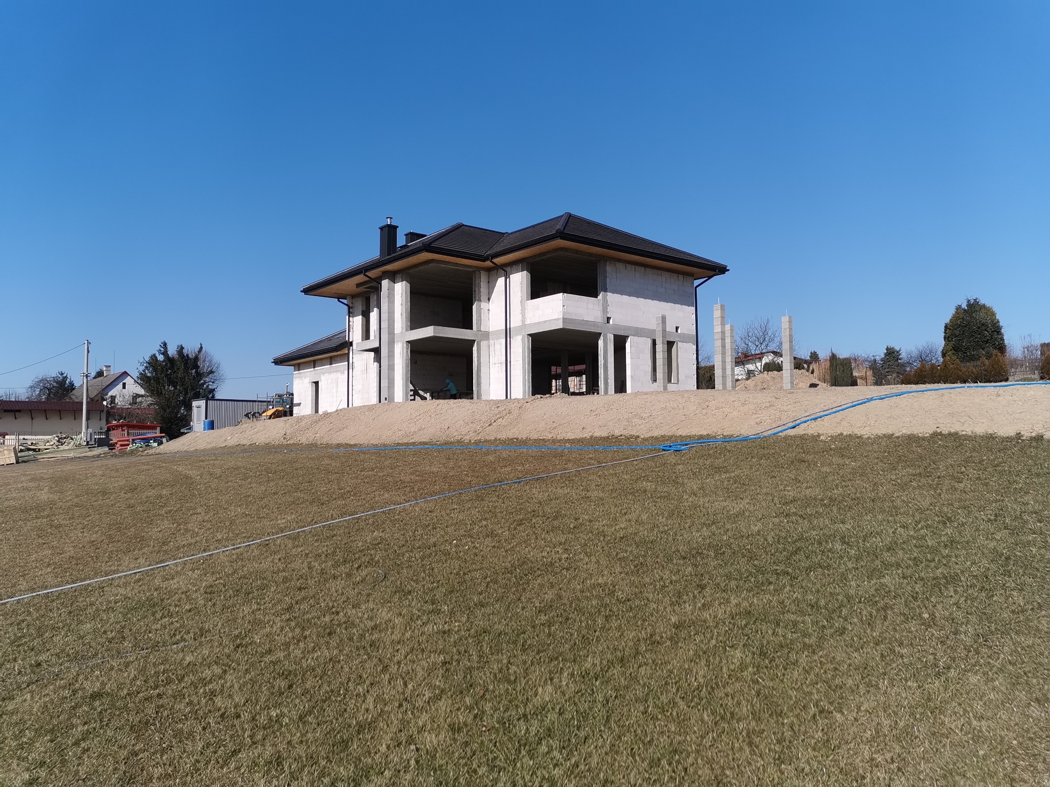 Dwukondygnacyjny dom w stanie surowym otwartym z czarnym dachem, wzniesiony na wzniesieniu, widok z dołu na tle błękitnego nieba.