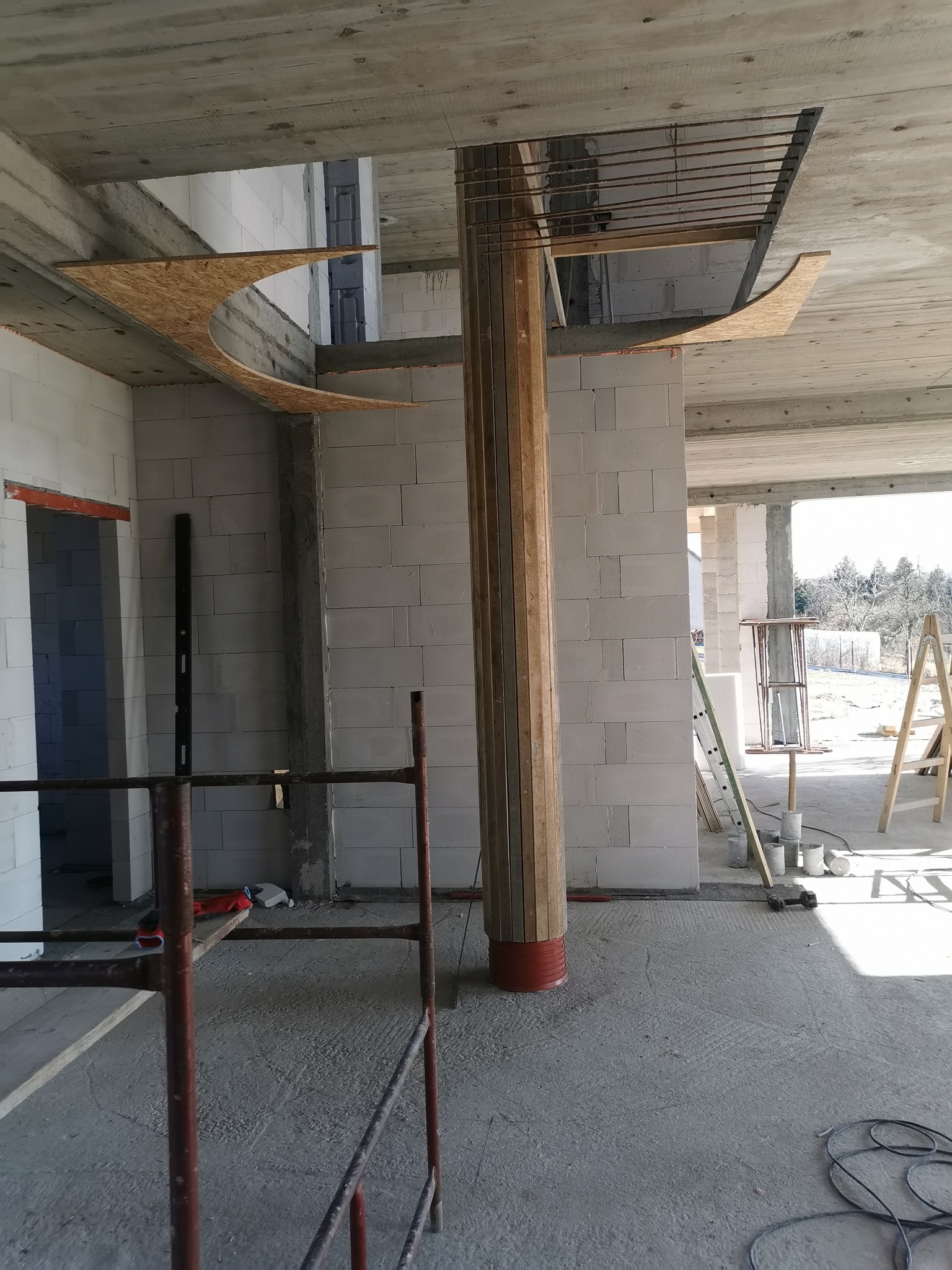 Wnętrze w stanie surowym: murowane ściany z betonu komórkowego, drewniana kolumna w szalunku, fragment deskowania na suficie, rusztowanie i drabiny w tle.