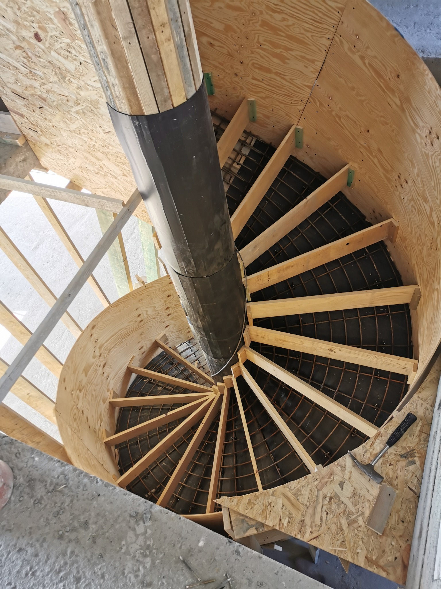 Widok z góry na szalunek spiralnych schodów z drewnianymi stopniami i centralnym słupem, zbrojenie wewnątrz konstrukcji, widoczny młotek ciesielski na płycie szalunkowej.