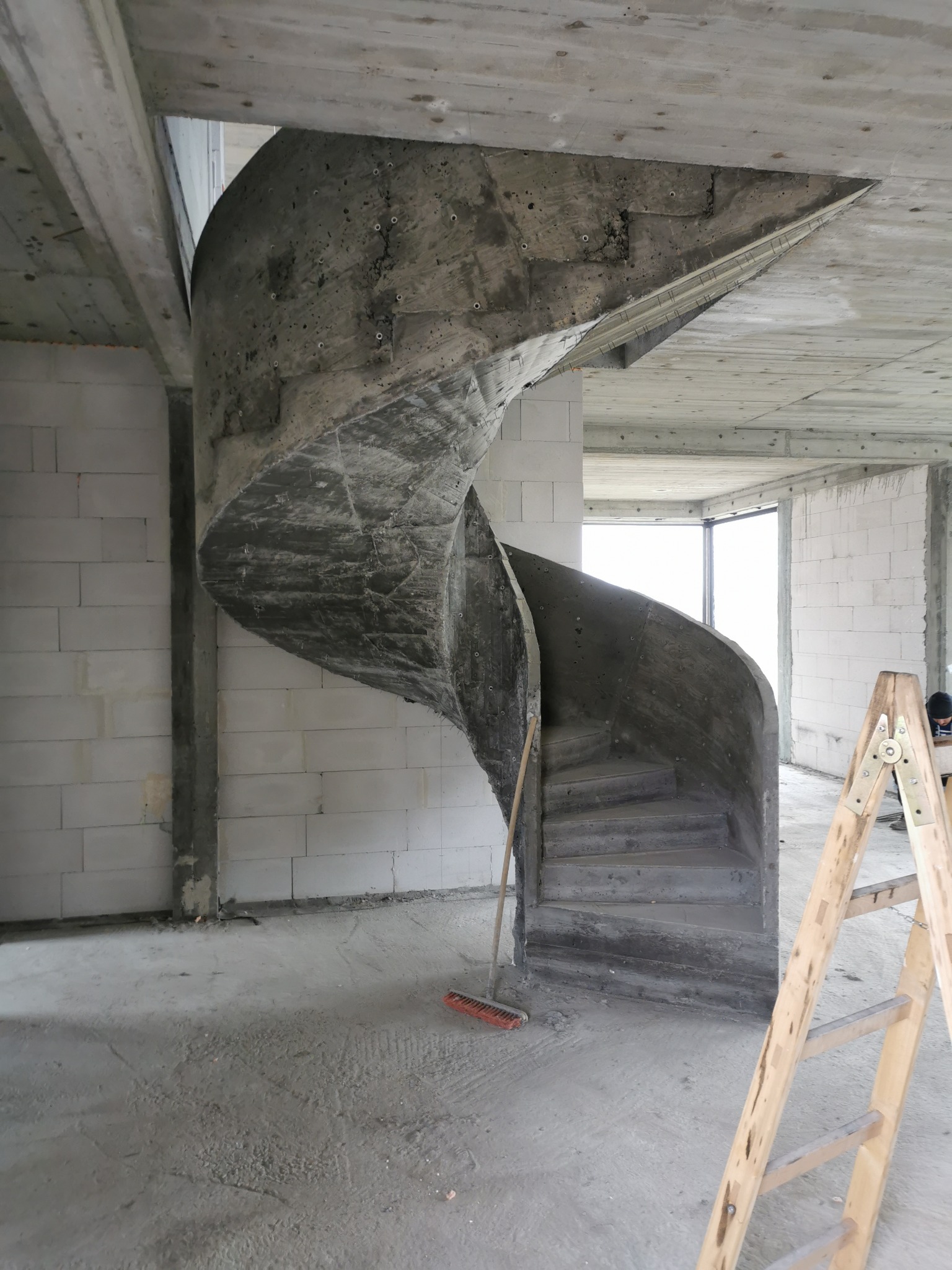 Surowe betonowe schody spiralne w surowym stanie, wewnątrz budynku w trakcie budowy, widoczna drabina drewniana i miotła.