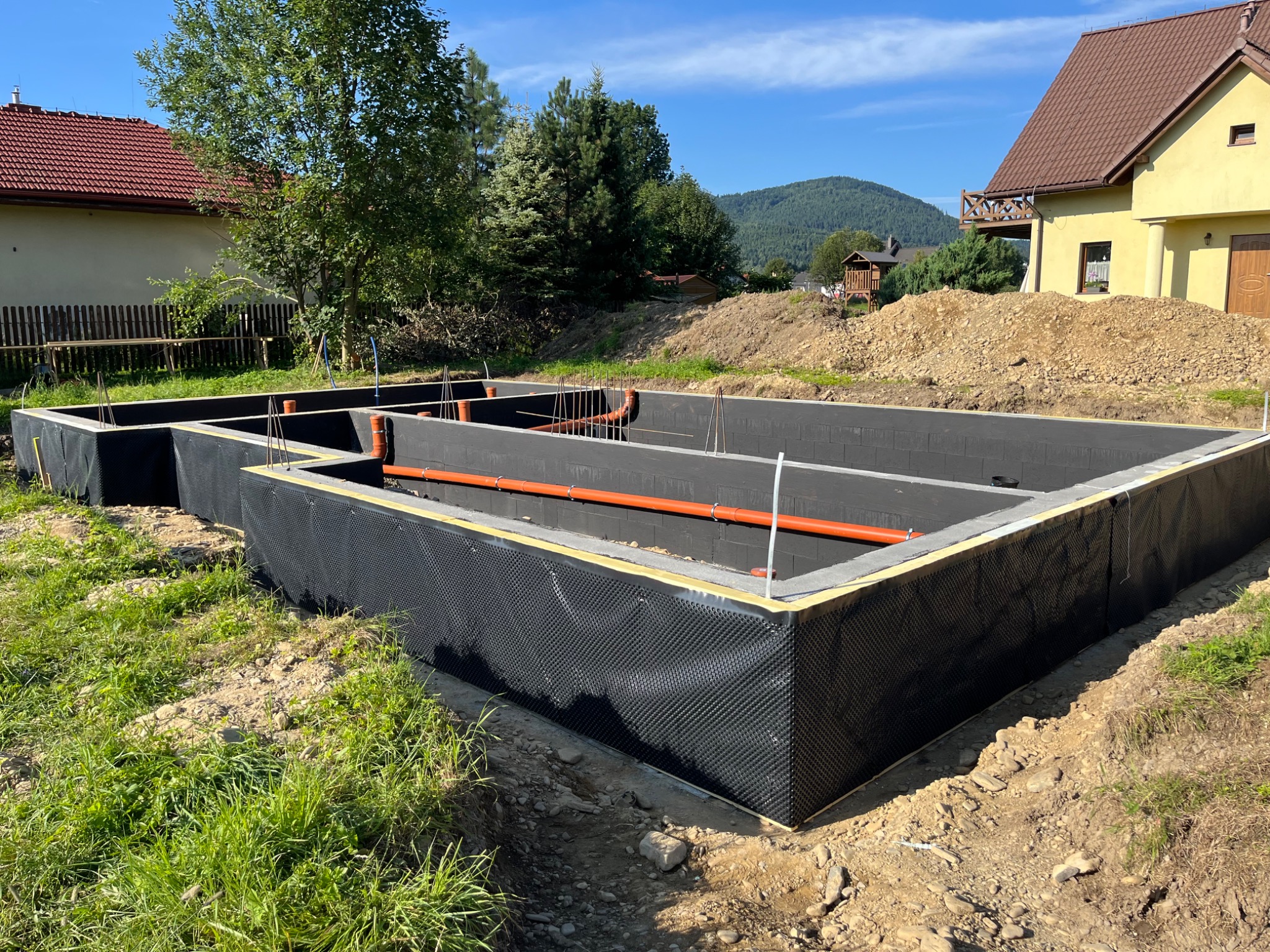 Zabezpieczone fundamenty z izolacją pionową i poziomą, wystające rury kanalizacyjne. W tle domy i drzewa w słoneczny dzień.