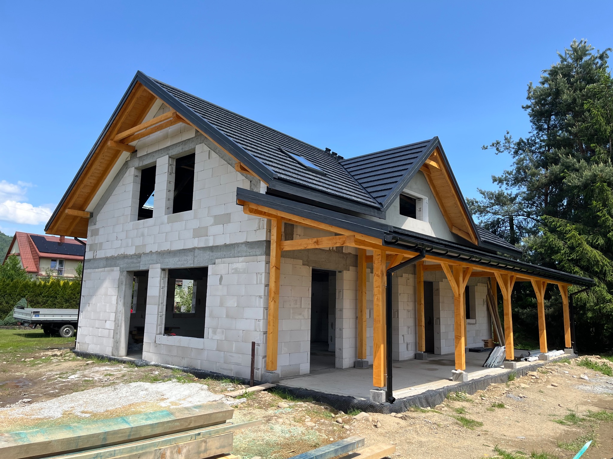 Surowy dom z białych bloczków, czarnym dachem i drewnianą konstrukcją werandy w słoneczny dzień. Widoczne niezabudowane otwory okienne i drzwiowe.