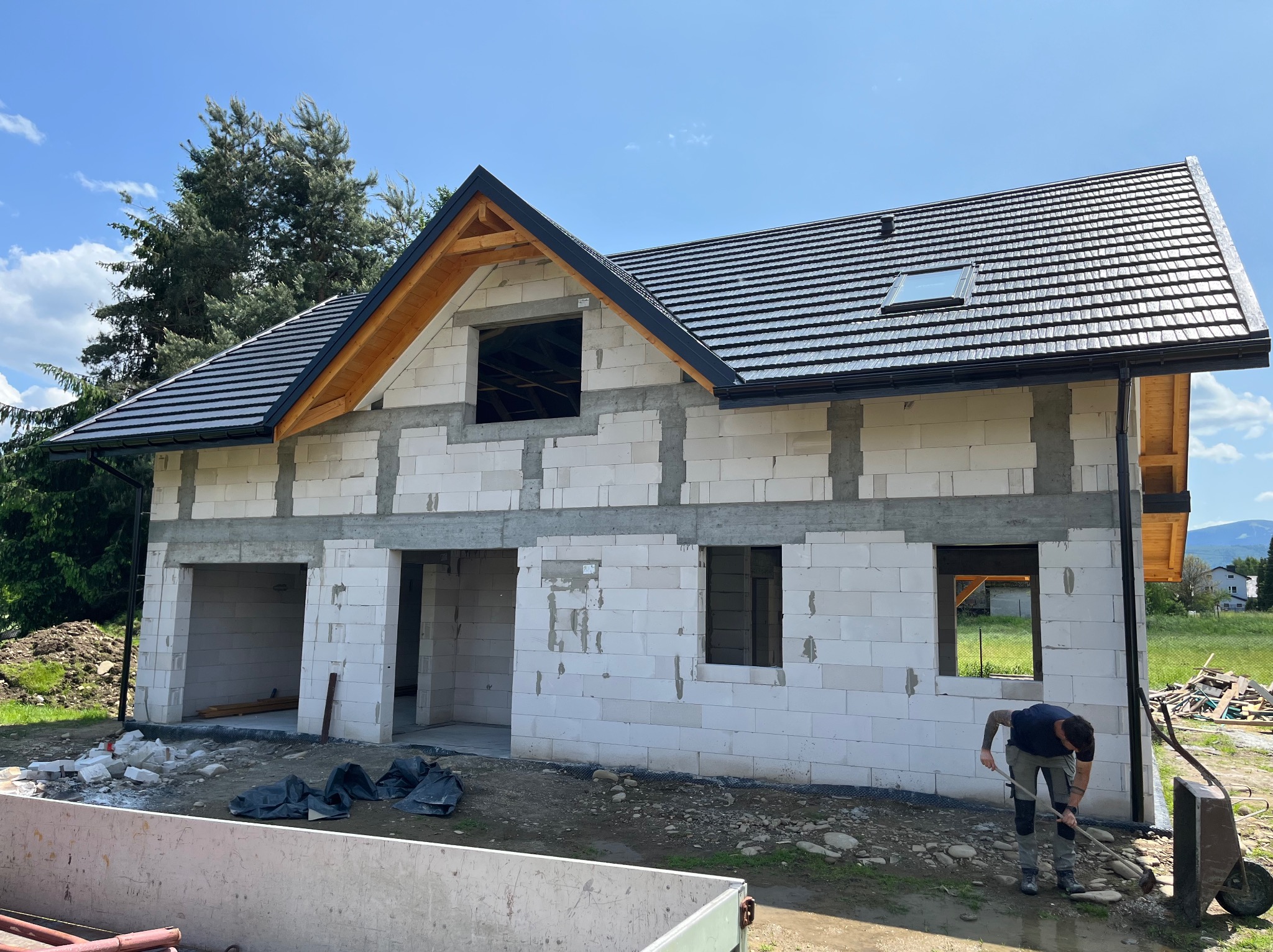 Surowy stan domu z białych bloczków, czarny dach z lukarną, pracownik na zewnątrz. Widok na góry w tle. Dom w trakcie budowy.
