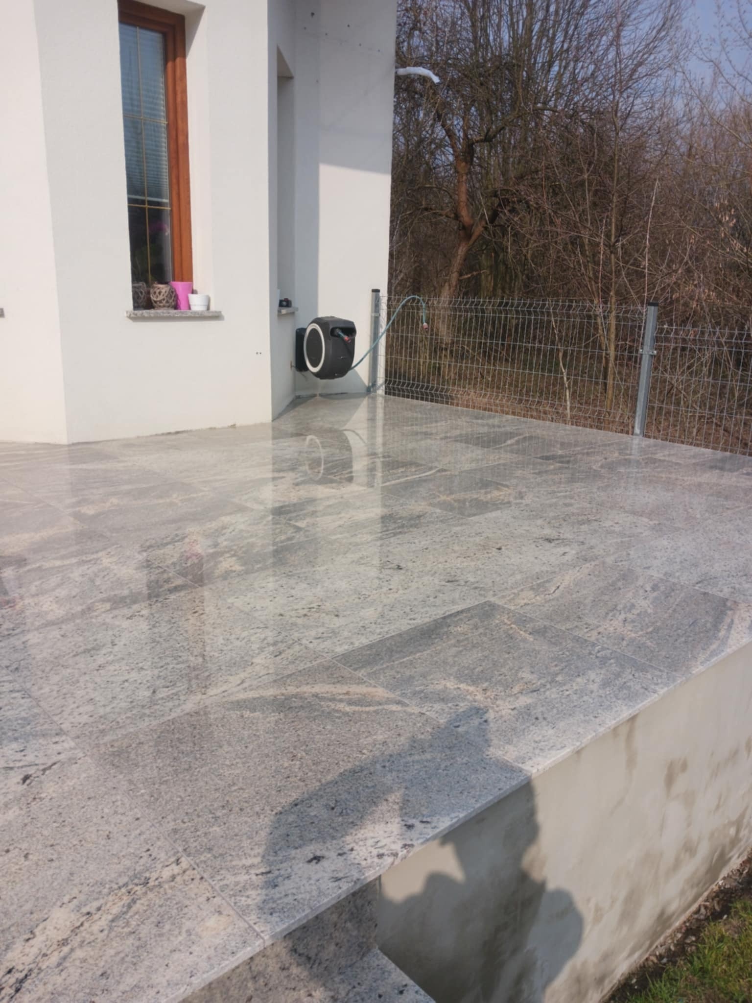 Wykończony taras z jasnoszarej, polerowanej płyty granitowej, przylegający do budynku z oknem. Widoczna zwinięta lina ogrodowa i siatka ogrodzeniowa w tle.