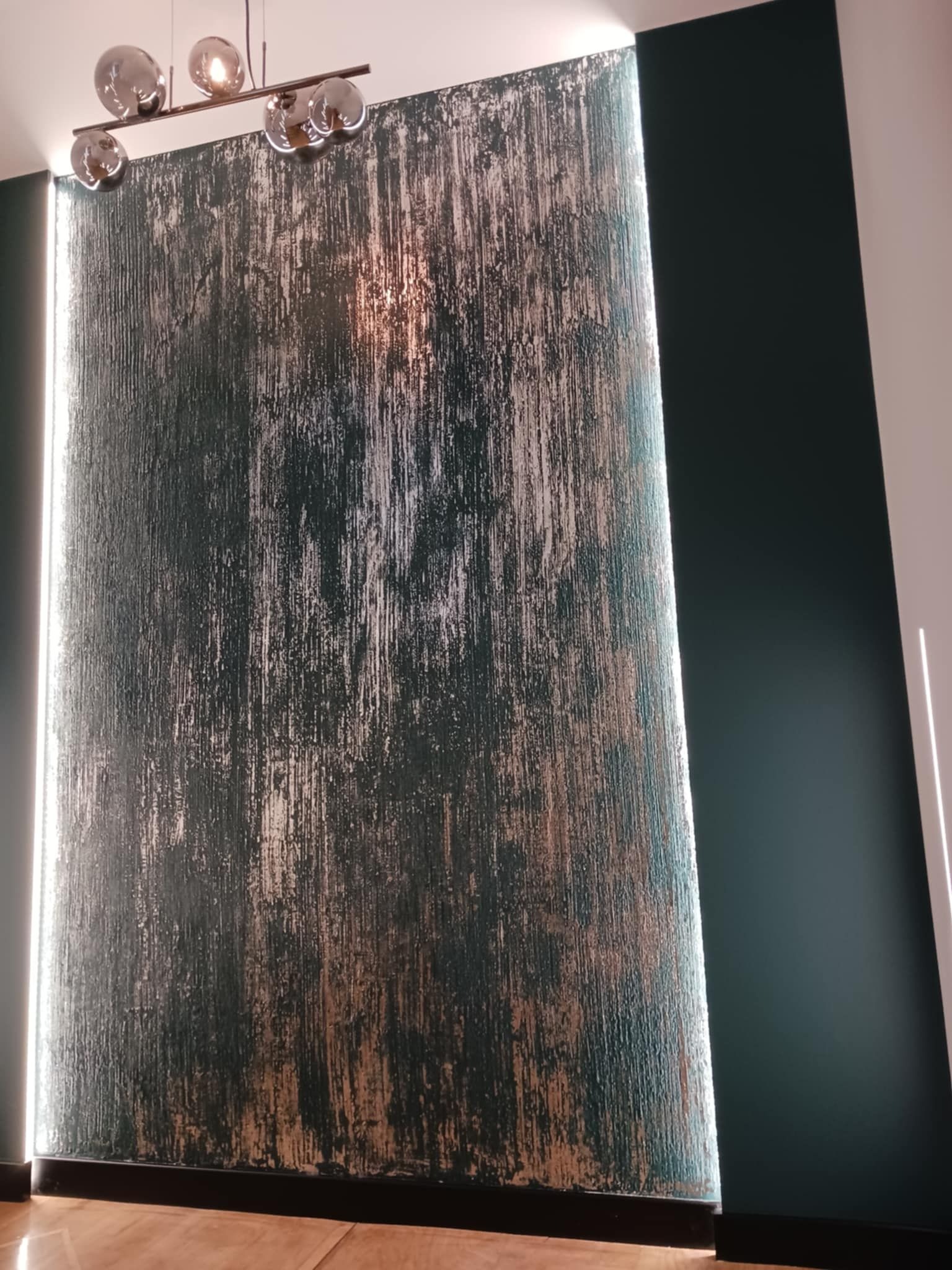 Dekoracyjna ściana z efektem przetarć w odcieniach zieleni i złota, oświetlona listwami LED. Nowoczesne oświetlenie sufitowe z kulistymi kloszami.