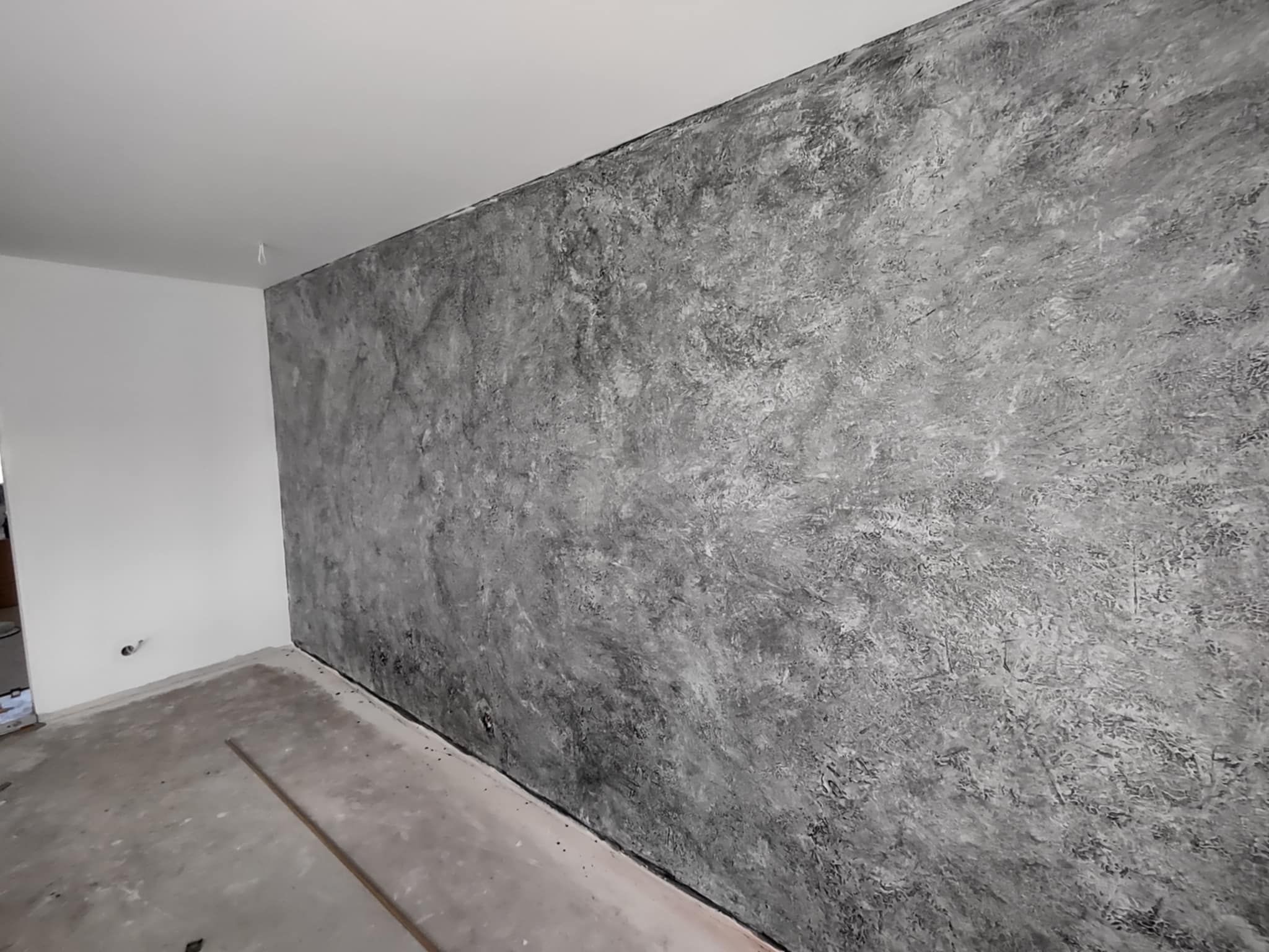 Ściana z efektem dekoracyjnym w odcieniach szarości, imitującym beton. Widoczne niedoskonałości i faktura. Surowy, nowoczesny styl wykończenia wnętrza. Częściowo wykończona podłoga.