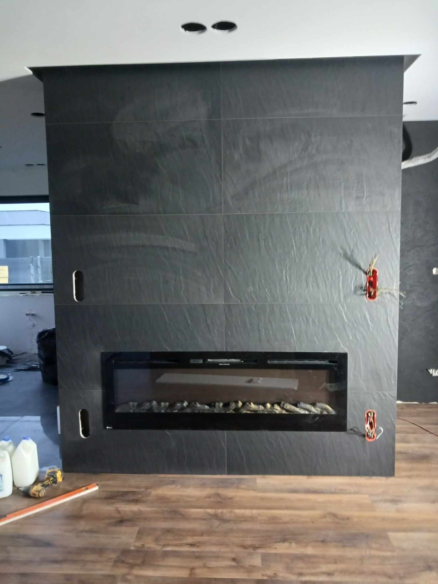 Ściana z szarych płytek imitujących kamień, z wbudowanym kominkiem elektrycznym. Widoczne puszki elektryczne i fragment podłogi z paneli.