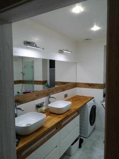 Nowoczesna łazienka w Wieliszewie z dwoma umywalkami na drewnianym blacie, dużym lustrem i pralką. Białe ściany z drewnianym akcentem. Oświetlenie punktowe.