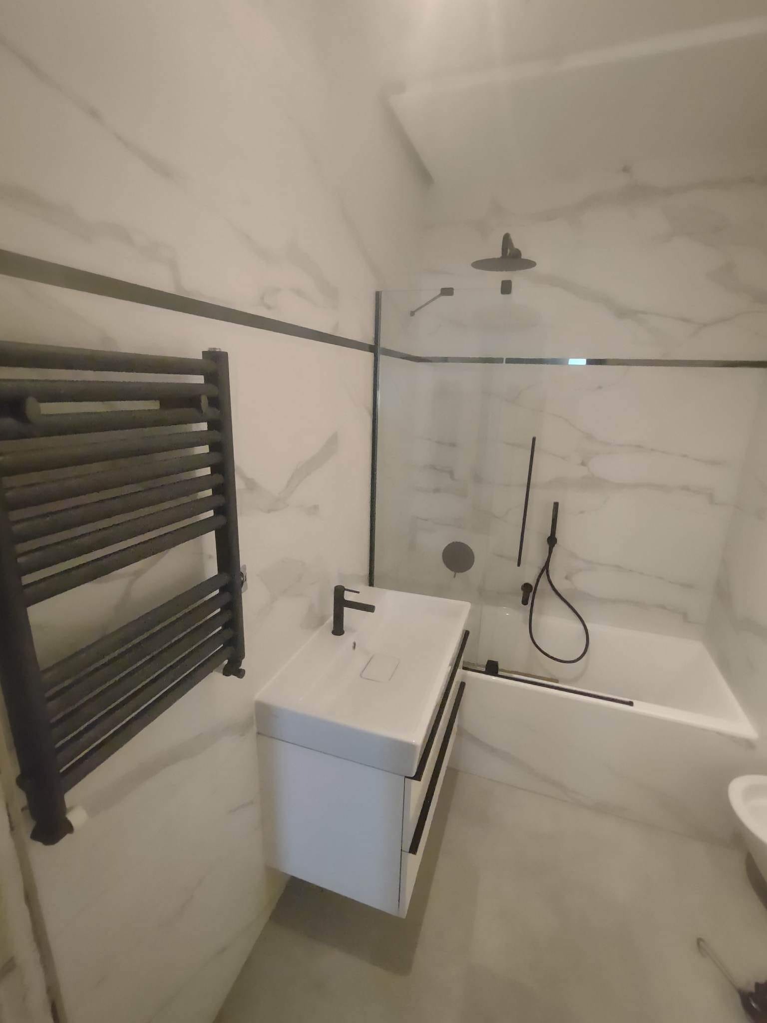 Nowoczesna łazienka z białymi marmurowymi płytkami, czarną armaturą i grzejnikiem drabinkowym. Minimalistyczny design, kabina prysznicowa ze szklaną ścianką.