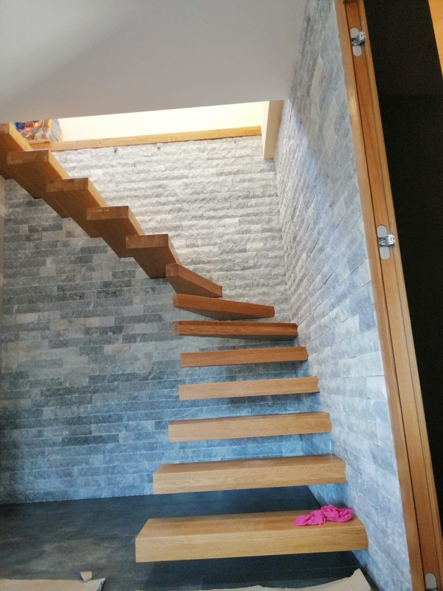 Nowoczesne, drewniane schody wiszące na tle ściany z kamienia dekoracyjnego. Widoczny fragment drzwi z ukrytymi zawiasami. Minimalistyczny design.