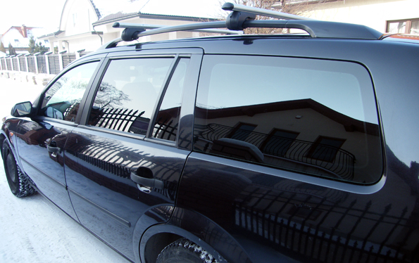 Ciemny samochód kombi z przyciemnionymi szybami, zaparkowany na śniegu w słoneczny dzień; w odbiciu na szybach widać budynek mieszkalny z balkonem.