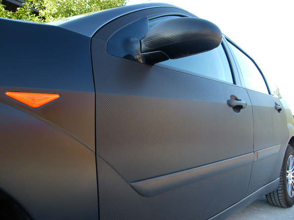 Samochód oklejony czarną folią imitującą karbon, widoczne detale struktury folii na karoserii, lusterku i klamkach.