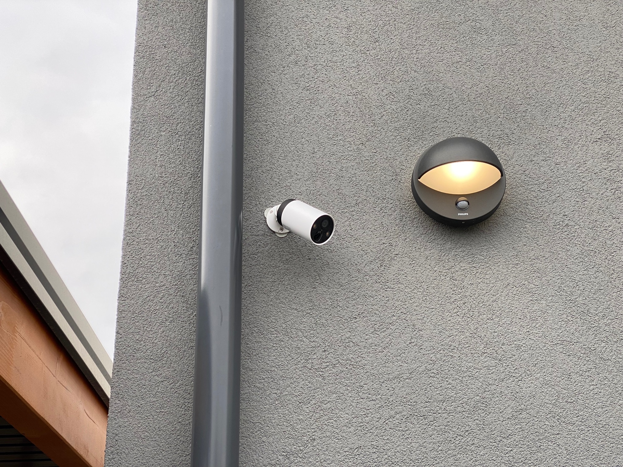 Biała kamera monitoringu na szarej elewacji domu, obok lampa Philips. Widoczny fragment rury spustowej i drewnianej konstrukcji dachu. Dyskretny system zabezpieczeń.