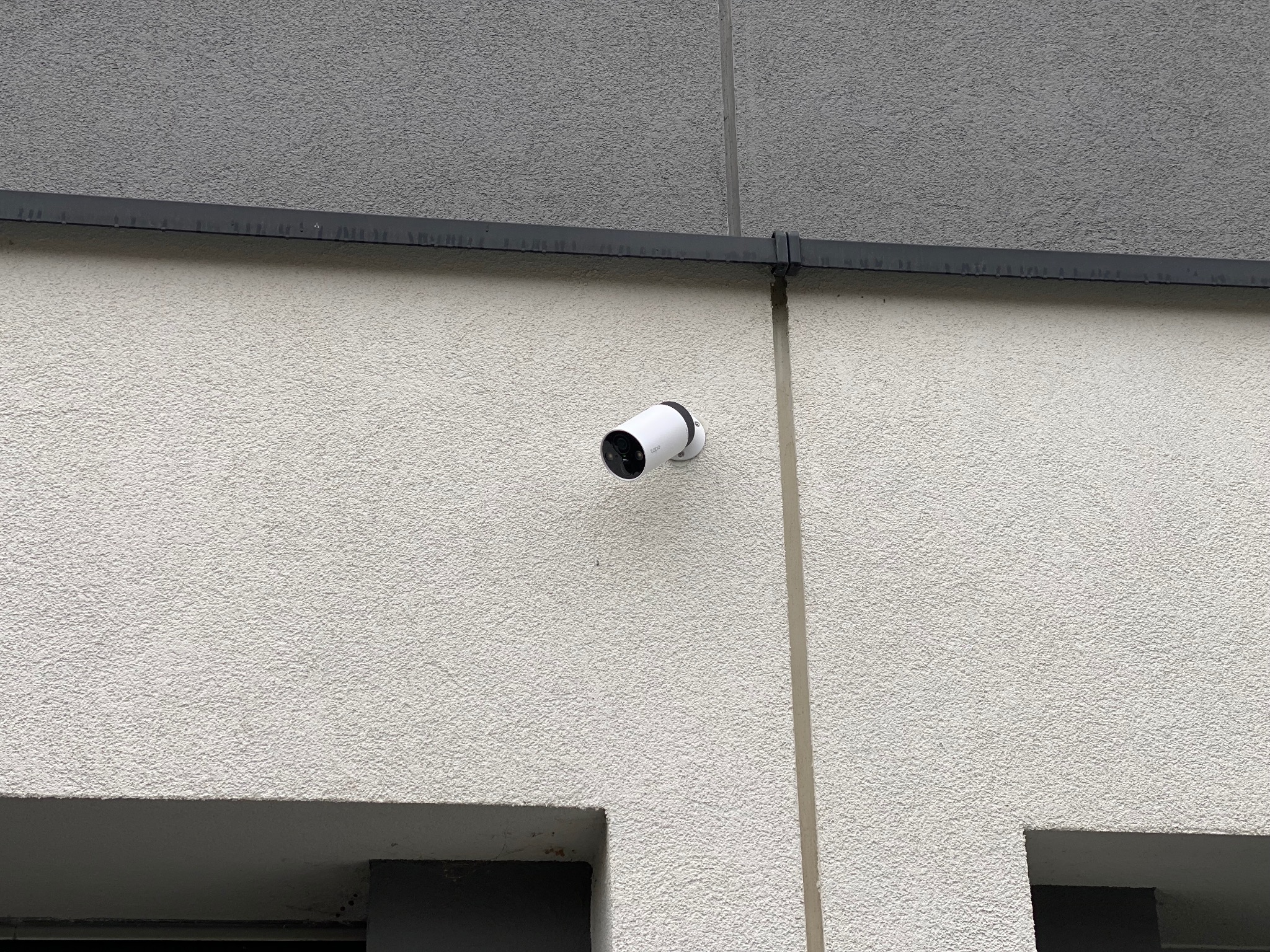 Biała kamera monitoringu zamontowana na elewacji budynku o jasnej strukturze, widoczna rynna. Ujęcie z dołu, podkreślające dyskretny charakter instalacji.