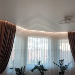 Nowoczesny salon z białym sufitem napinanym, oświetleniem LED wzdłuż krawędzi, brązowymi zasłonami i roślinami doniczkowymi przy oknie.