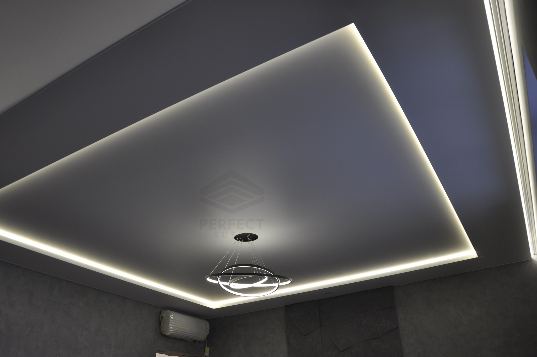 Szary, matowy sufit z wbudowanym oświetleniem LED w kształcie kwadratu i nowoczesnym, okrągłym żyrandolem w centrum, minimalistyczny design.