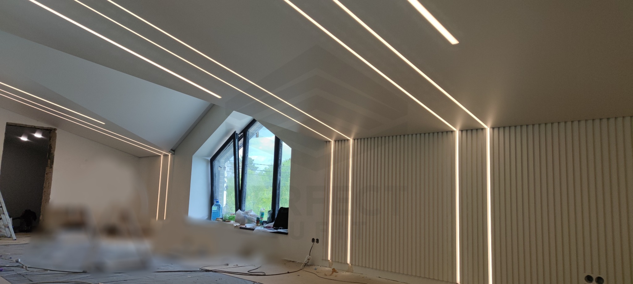 Wnętrze poddasza z oknem, wykończone panelami ściennymi i oświetleniem liniowym LED w suficie i ścianach, widoczne ślady prac wykończeniowych na podłodze.