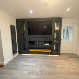 Wojciech Słotwiński Ładniej Meble - Nowoczesna ściana telewizyjna z pionowymi panelami, wbudowanym elektrycznym kominkiem i półkami, na podłodze panele imitujące drewno.