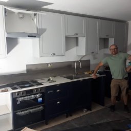 Wojciech Słotwiński Ładniej Meble - Montaż białych wiszących szafek kuchennych i ciemnych szafek dolnych z blatem, widoczny monter w okularach i roboczym ubraniu, częściowo zabezpieczony sprzęt AGD, podłoga w trakcie prac...