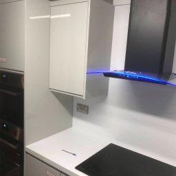 Wojciech Słotwiński Ładniej Meble - Nowoczesna zabudowa kuchenna z białymi, lakierowanymi frontami, piekarnikiem w zabudowie, czarnym okapem z niebieskim podświetleniem LED i blatem z delikatnym wzorem, na którym leży długopis.