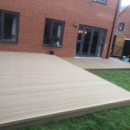 Wojciech Słotwiński Ładniej Meble - Beżowy taras kompozytowy przylegający do ceglanego domu z szarymi oknami i drzwiami tarasowymi, widok na zadbany trawnik.