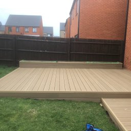 Wojciech Słotwiński Ładniej Meble - Kompozytowy taras w ogrodzie, czyszczony przez kobietę z pomarańczową szczotką, tło stanowią nowoczesne domy jednorodzinne i brązowe ogrodzenie.