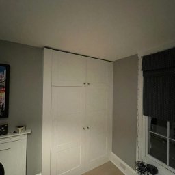 Wojciech Słotwiński Ładniej Meble - Biała, czterodrzwiowa szafa sięgająca sufitu, z małymi, okrągłymi uchwytami, umieszczona w rogu pokoju z szarymi ścianami i jasną podłogą, obok okna z zasłoniętą roletą rzymską.