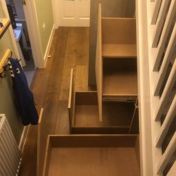 Wojciech Słotwiński Ładniej Meble - Otwarta szafa wnękowa pod schodami z wysuwanymi szufladami, widok z góry na drewnianą podłogę i białą balustradę.