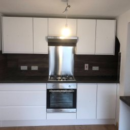 Wojciech Słotwiński Ładniej Meble - Nowoczesna kuchnia z białymi szafkami, ciemnym blatem i stalowym okapem, z piekarnikiem w folii ochronnej, pod wiszącą żarówką bez klosza.