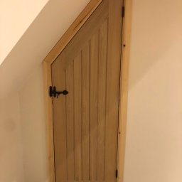 Wojciech Słotwiński Ładniej Meble - Dopasowane drewniane drzwi do wnęki pod skosem, z czarnym kutym zamkiem i pionowymi panelami, osadzone w drewnianej ramie.