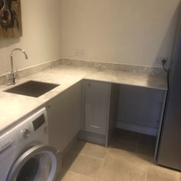 Wojciech Słotwiński Ładniej Meble - Szara zabudowa kuchenna z blatem imitującym marmur, zintegrowany zlewozmywak, pralka Bosch z otwartymi drzwiami i obraz krowy na ścianie w tle.