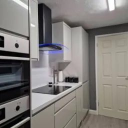 Nowoczesna kuchnia w odcieniach szarości z błyszczącymi frontami, czarną płytą indukcyjną i okapem z niebieskim podświetleniem LED oraz srebrnym stojakiem na akcesoria kuchenne na blacie.