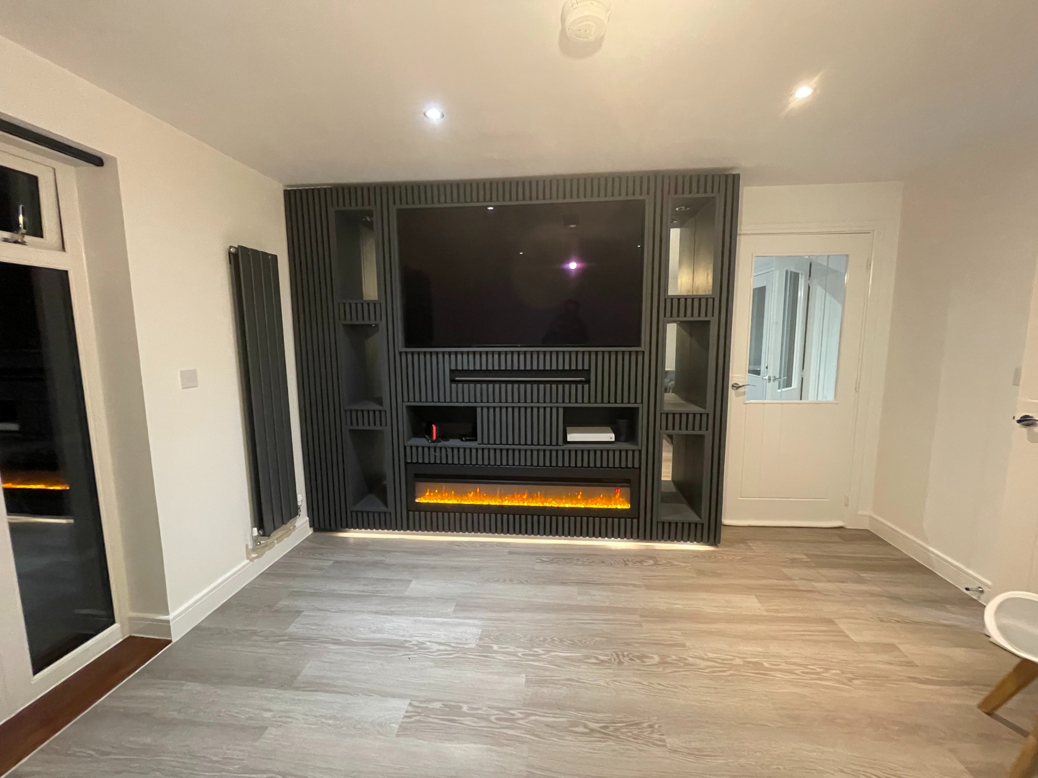 Nowoczesna ściana telewizyjna z pionowymi panelami, wbudowanym elektrycznym kominkiem i półkami, na podłodze panele imitujące drewno.