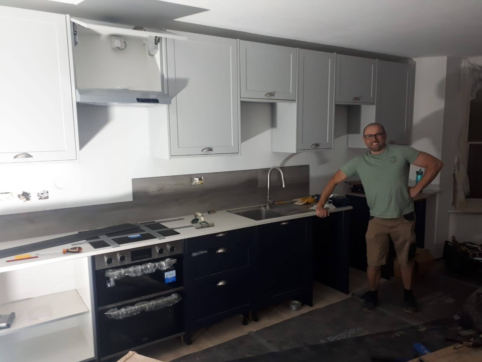 Montaż białych wiszących szafek kuchennych i ciemnych szafek dolnych z blatem, widoczny monter w okularach i roboczym ubraniu, częściowo zabezpieczony sprzęt AGD, podłoga w trakcie prac...