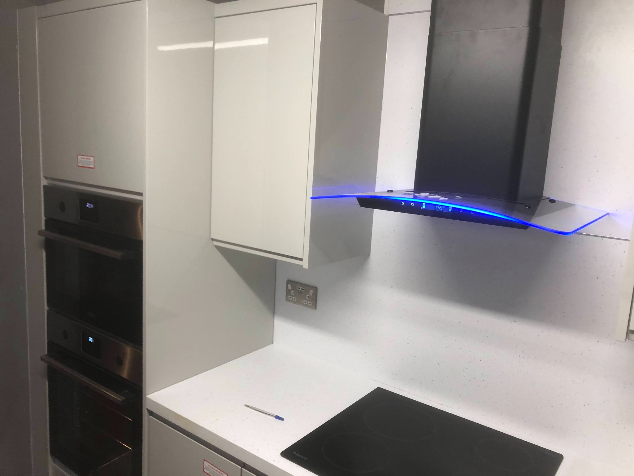 Nowoczesna zabudowa kuchenna z białymi, lakierowanymi frontami, piekarnikiem w zabudowie, czarnym okapem z niebieskim podświetleniem LED i blatem z delikatnym wzorem, na którym leży długopis.