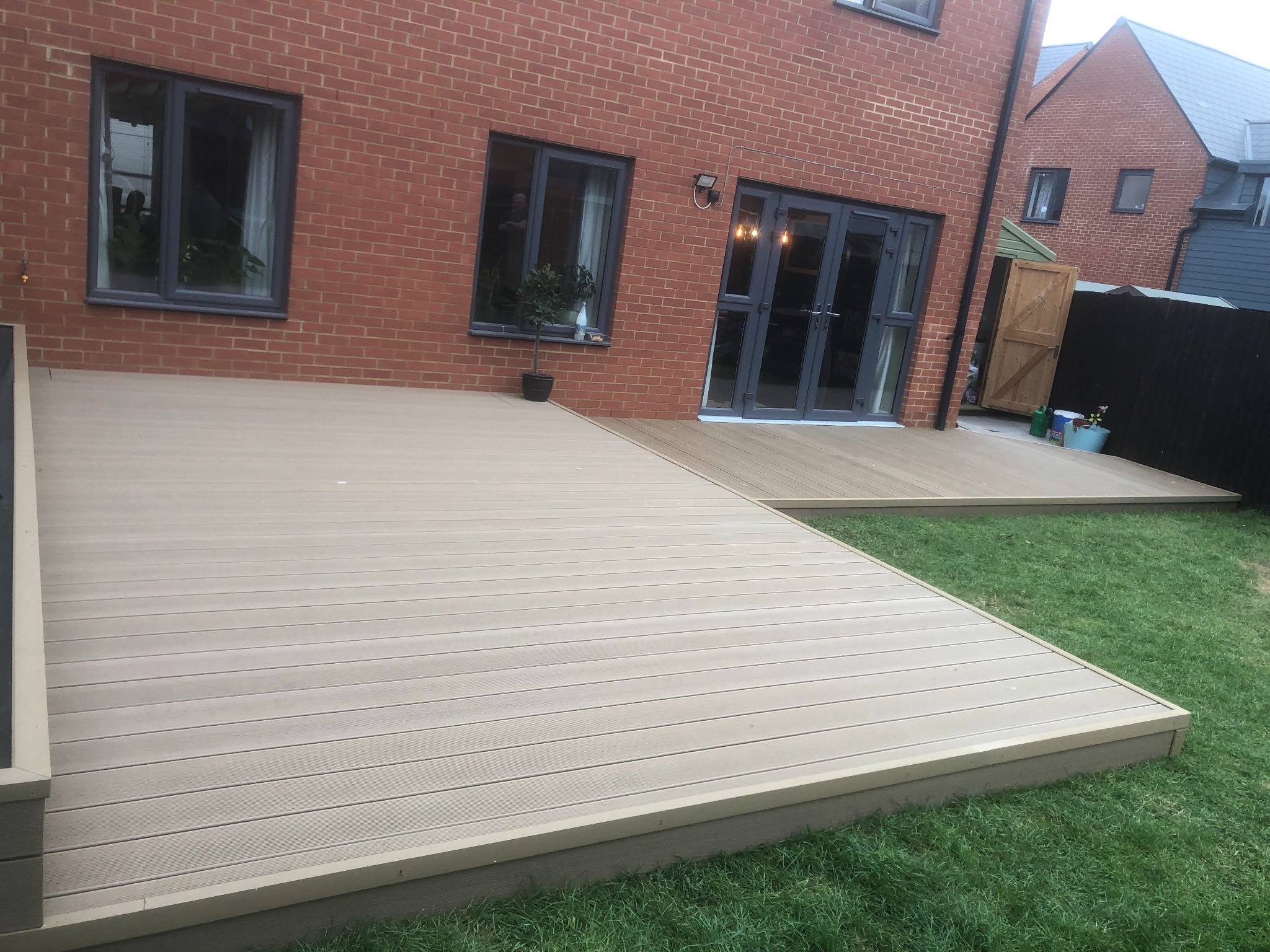 Beżowy taras kompozytowy przylegający do ceglanego domu z szarymi oknami i drzwiami tarasowymi, widok na zadbany trawnik.