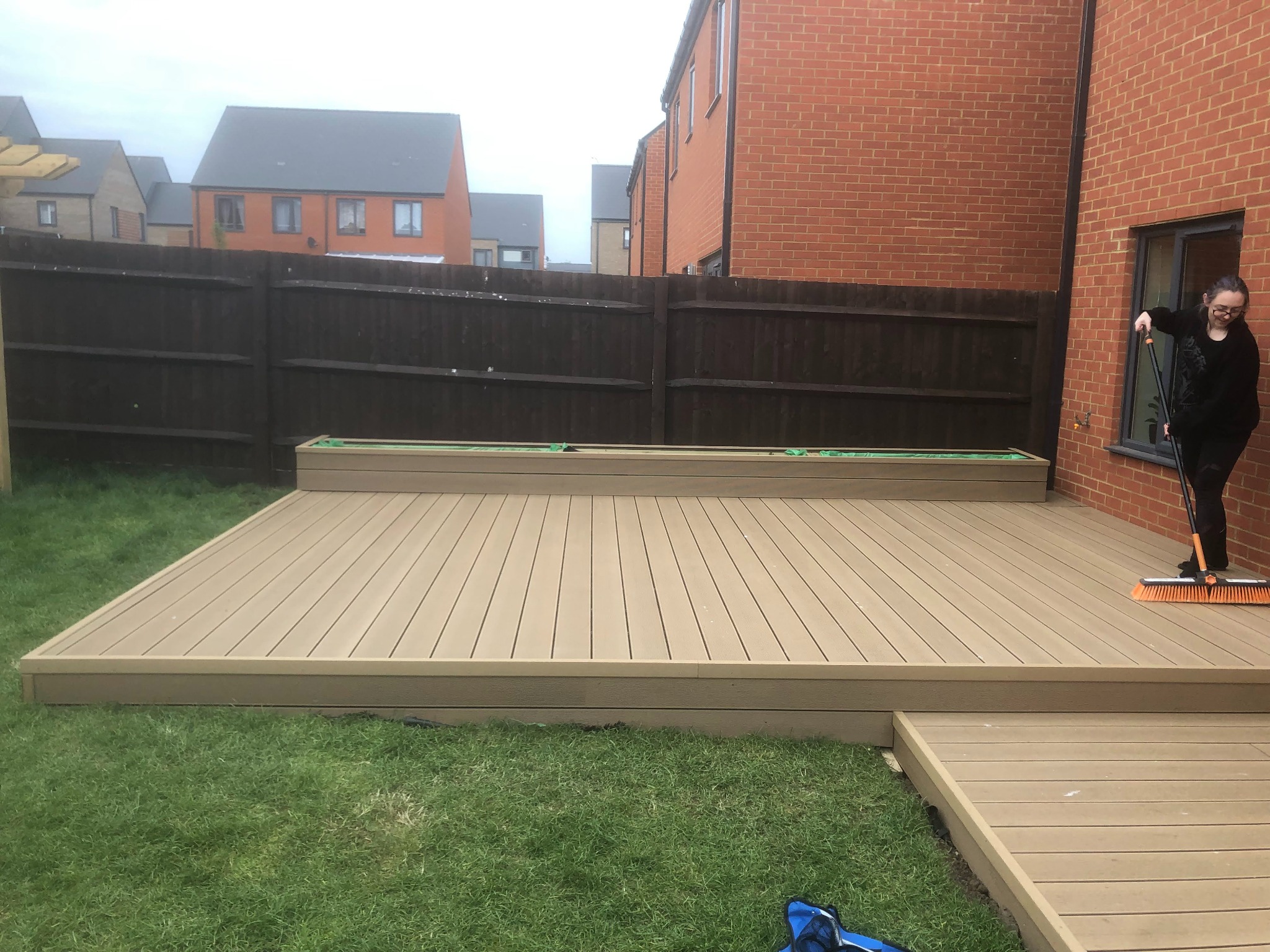 Kompozytowy taras w ogrodzie, czyszczony przez kobietę z pomarańczową szczotką, tło stanowią nowoczesne domy jednorodzinne i brązowe ogrodzenie.