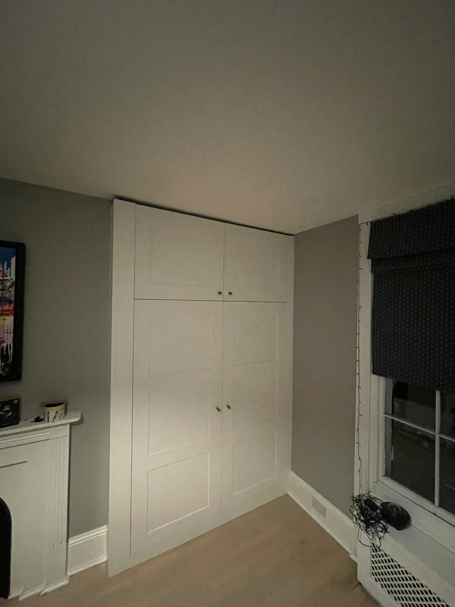 Biała, czterodrzwiowa szafa sięgająca sufitu, z małymi, okrągłymi uchwytami, umieszczona w rogu pokoju z szarymi ścianami i jasną podłogą, obok okna z zasłoniętą roletą rzymską.