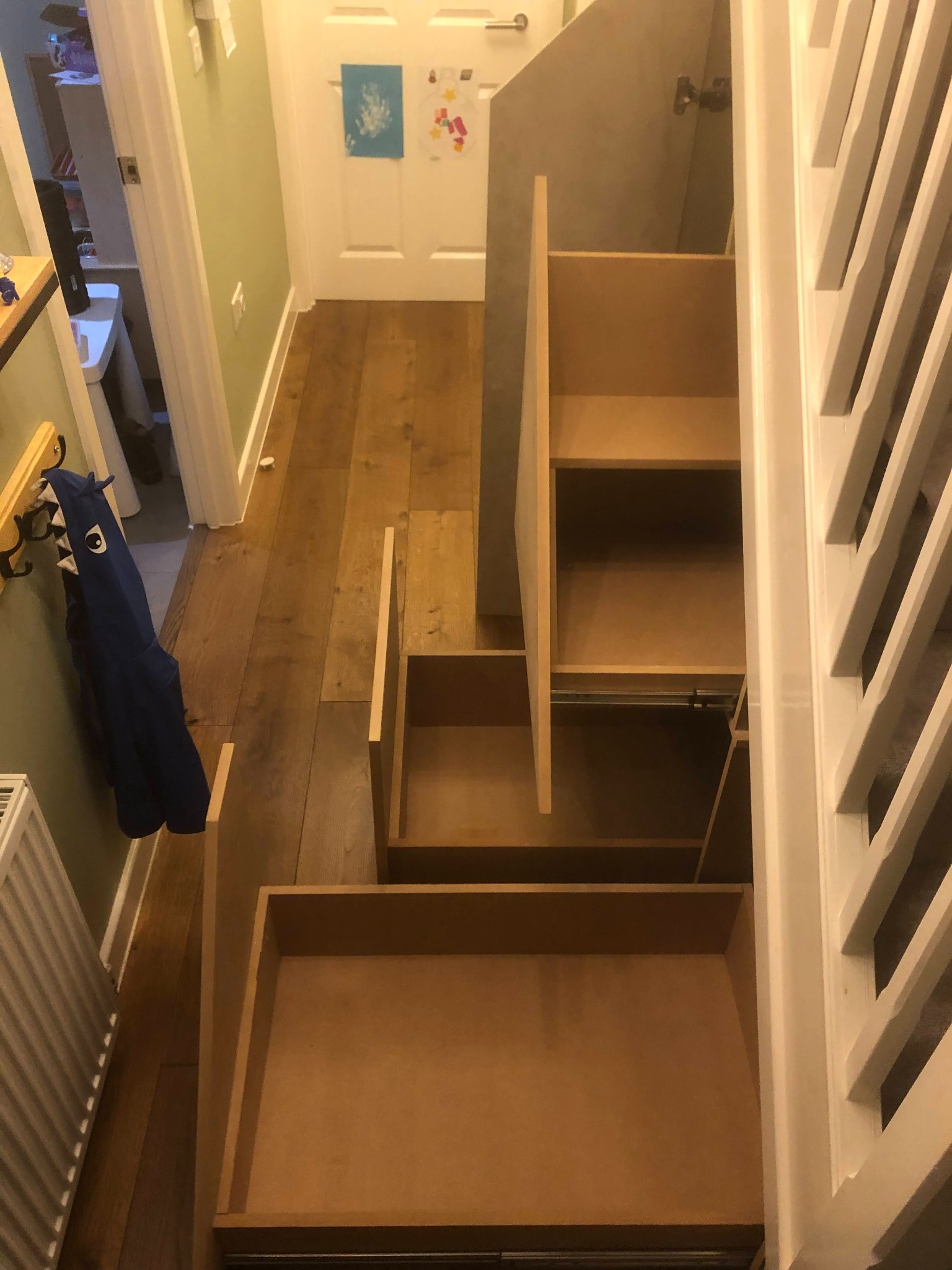 Otwarta szafa wnękowa pod schodami z wysuwanymi szufladami, widok z góry na drewnianą podłogę i białą balustradę.