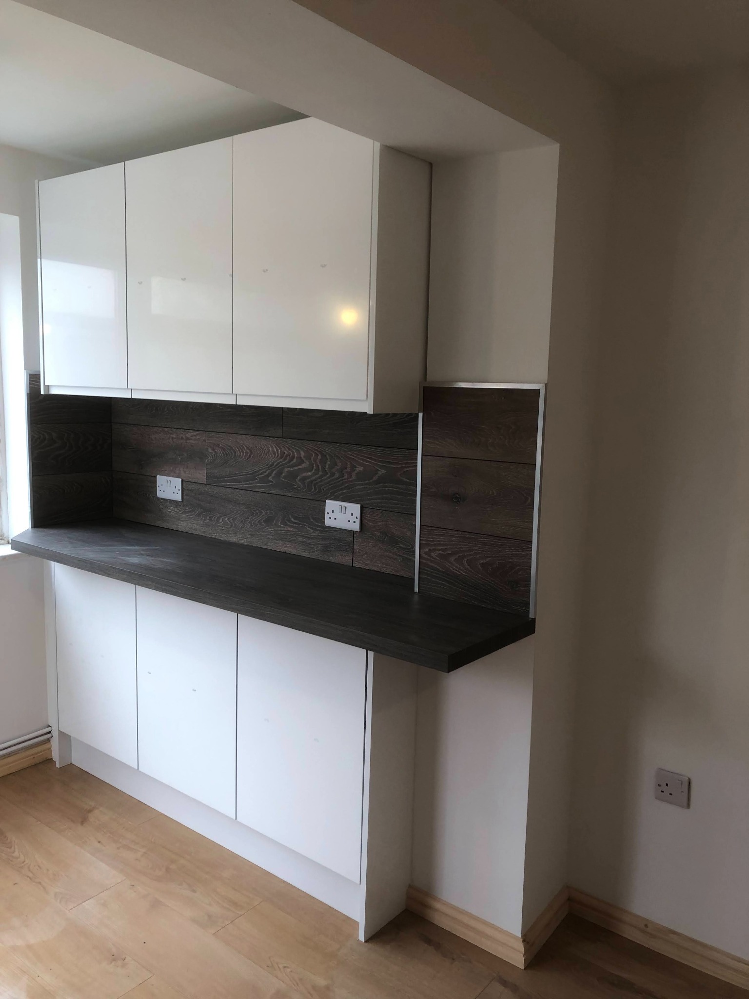 Białe, lakierowane szafki kuchenne z ciemnym blatem i drewnopodobnym panelem ściennym z gniazdkami elektrycznymi, widok z dołu.