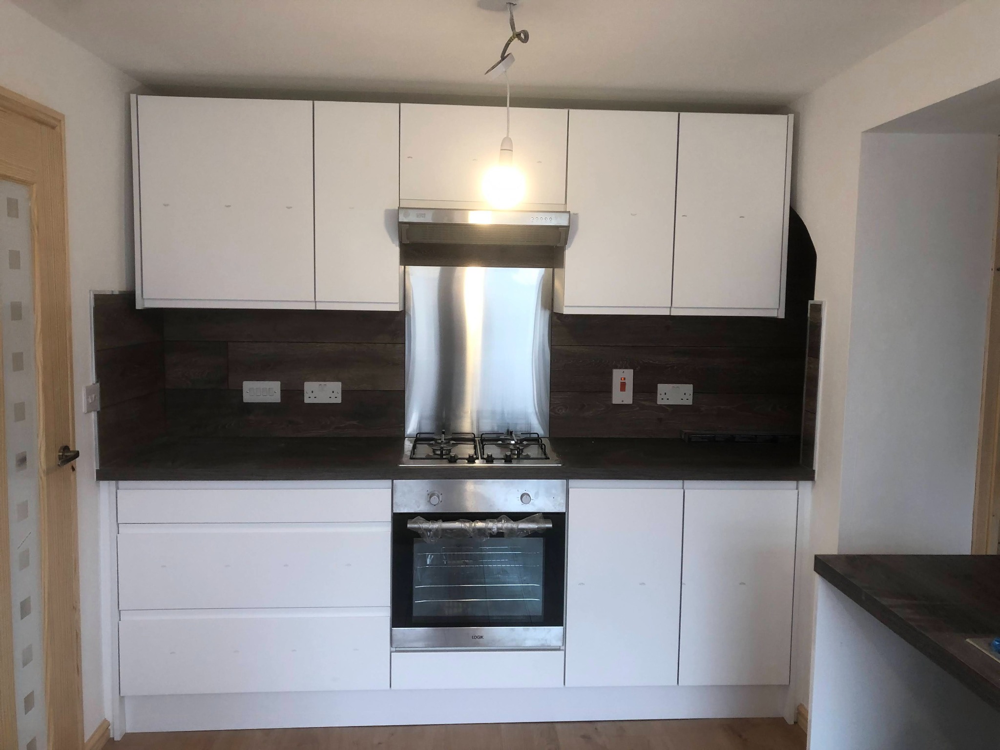 Nowoczesna kuchnia z białymi szafkami, ciemnym blatem i stalowym okapem, z piekarnikiem w folii ochronnej, pod wiszącą żarówką bez klosza.