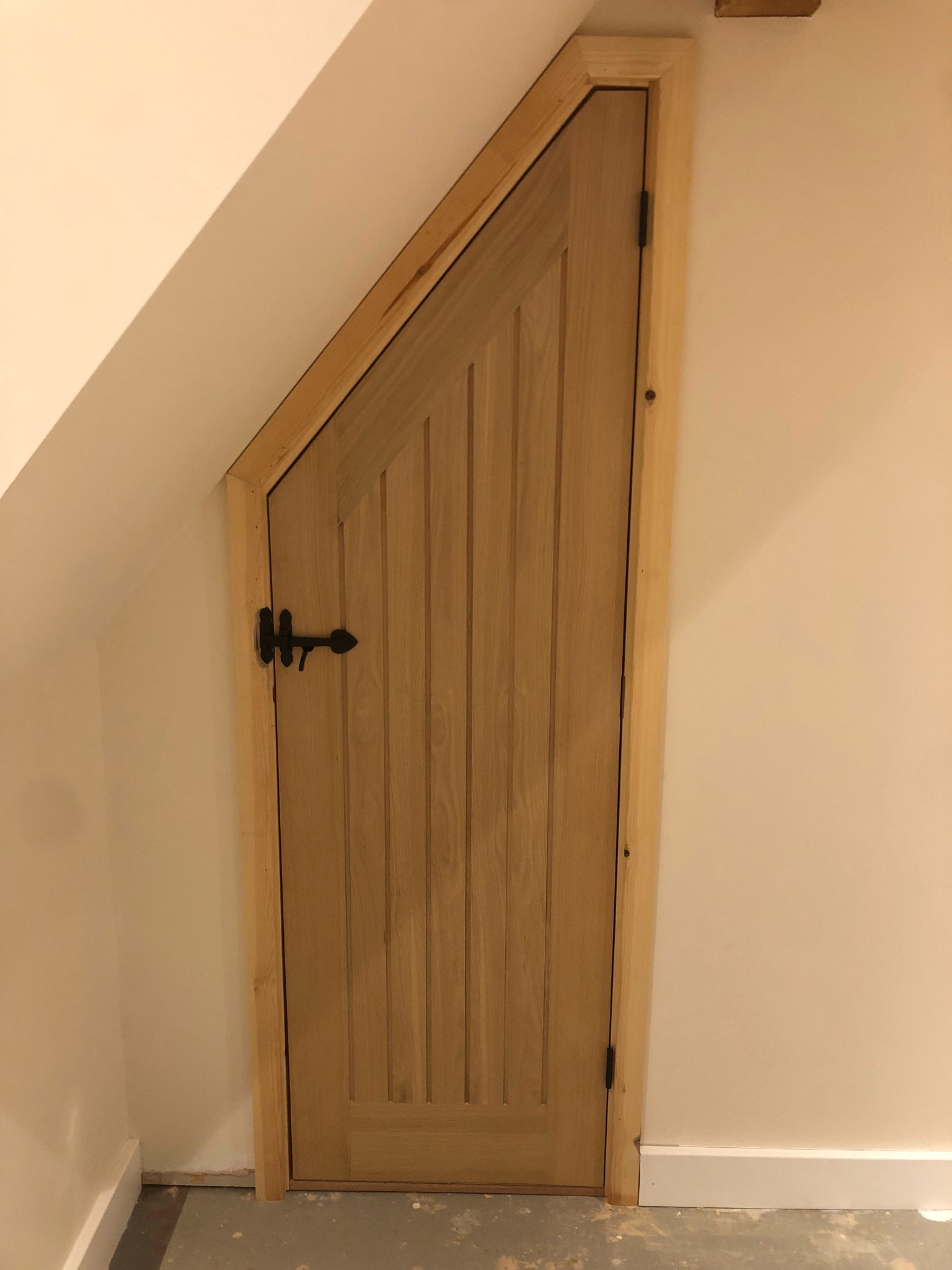 Dopasowane drewniane drzwi do wnęki pod skosem, z czarnym kutym zamkiem i pionowymi panelami, osadzone w drewnianej ramie.