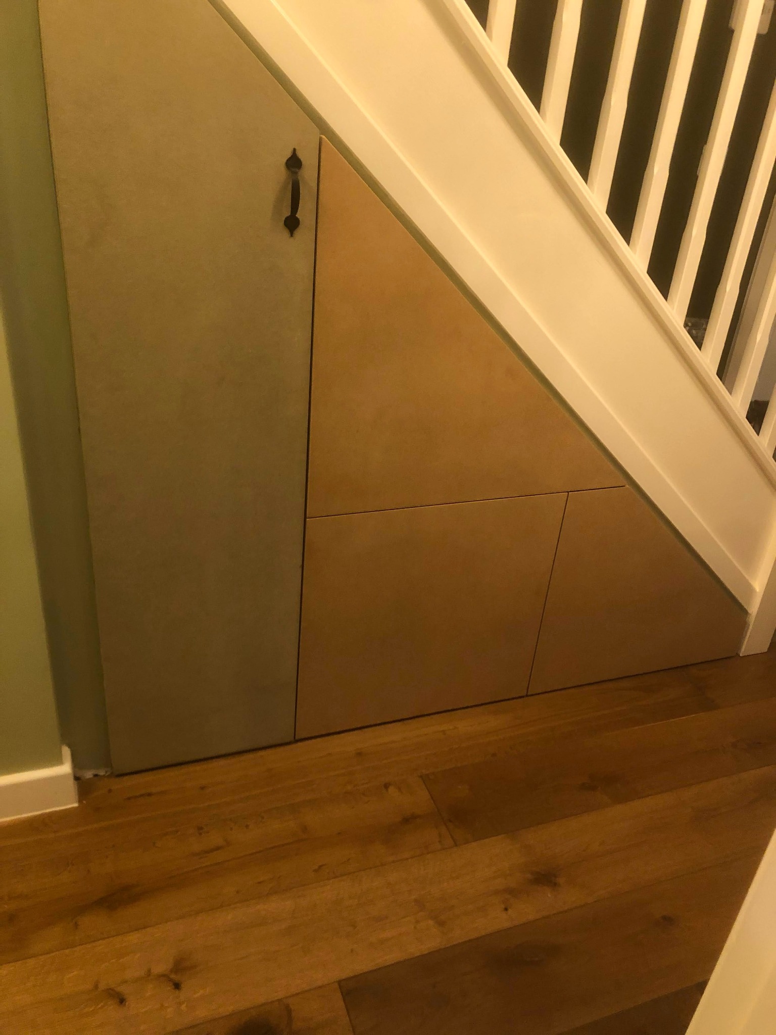 Zabudowa pod schodami z czterema drzwiczkami, z prostym uchwytem na jednych z nich, na drewnianej podłodze, widok na białą balustradę schodów.