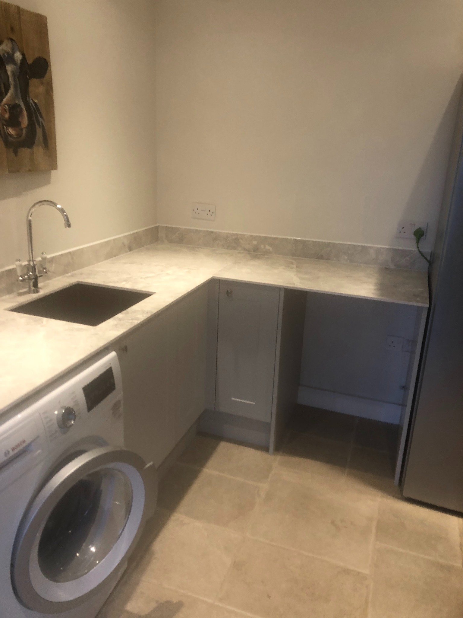 Szara zabudowa kuchenna z blatem imitującym marmur, zintegrowany zlewozmywak, pralka Bosch z otwartymi drzwiami i obraz krowy na ścianie w tle.