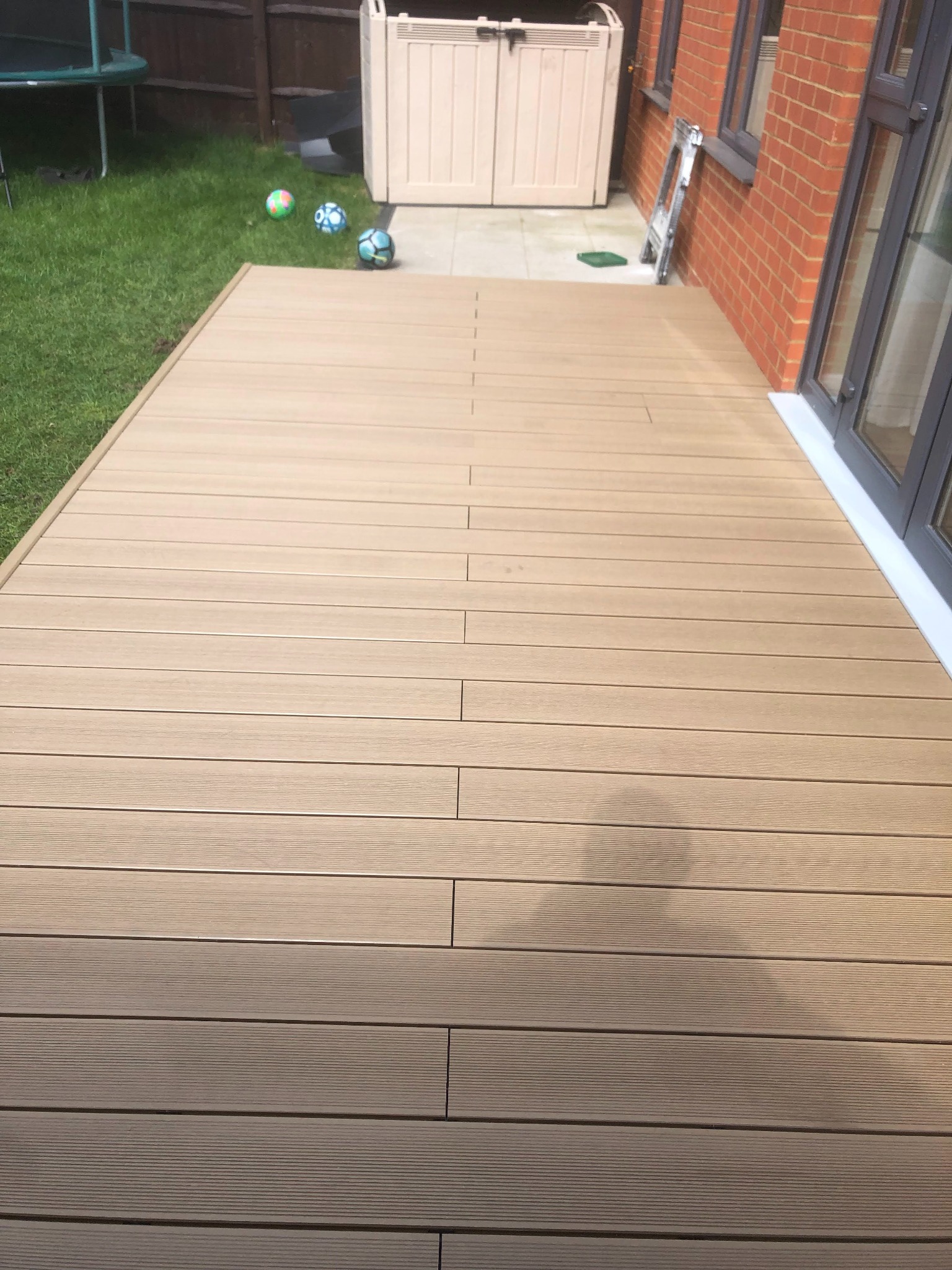 Nowoczesny taras z desek kompozytowych w odcieniach beżu, przylegający do domu z cegły, z widocznym trawnikiem, trampoliną i skrzynią ogrodową w tle.