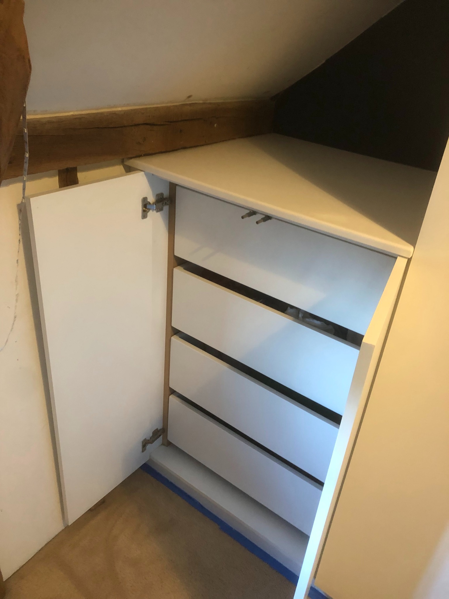 Biała szafa z szufladami we wnęce na poddaszu, otwarte drzwiczki po lewej stronie, widoczne drewniane belki stropowe i zabezpieczenie podłogi taśmą malarską.